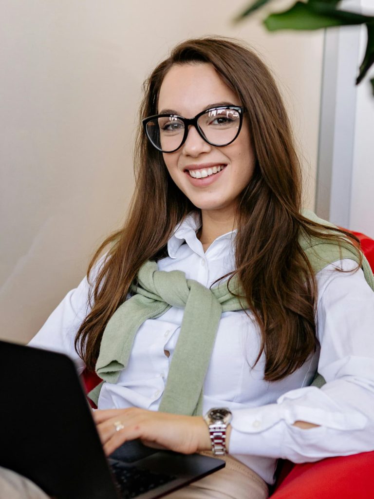 Mujer jóven con lentes y su laptop sentada en un sillón sonriendo
