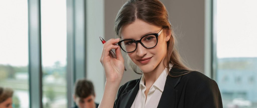 Mujer jóven profesionista con lentes y bolígrafo en un despacho corporativo contenta de ayudar a sus clientes