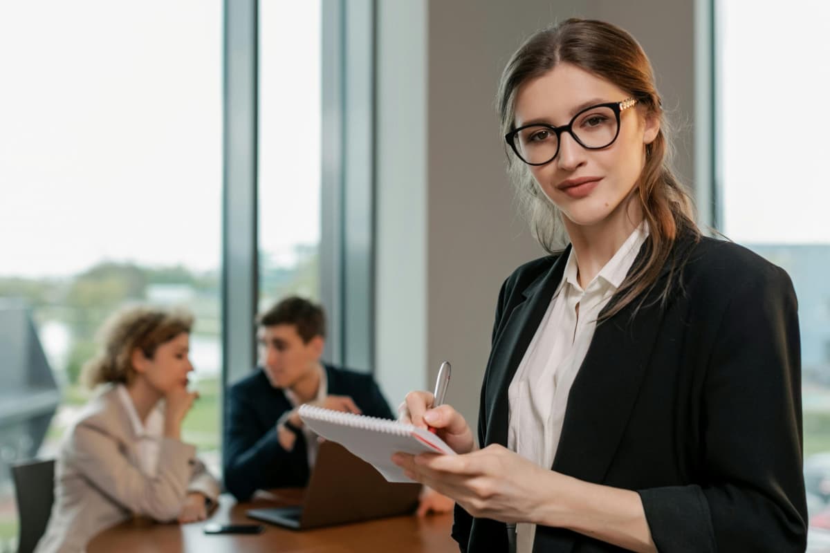 Mujer joven profesionista con lentes tomando notas en su despacho con clientes